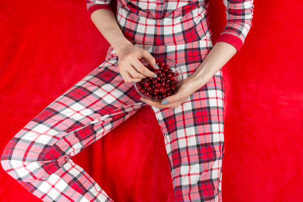 a woman in red checked home suite eats berries at home on a red background in quarantine coronavirus covid-19, without face - Foto, Imagen