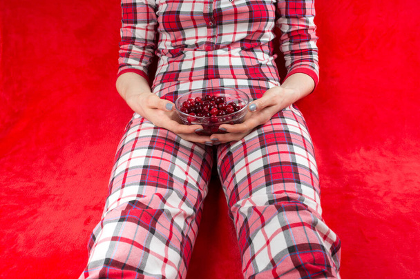 a woman in red checked home suite eats berries at home on a red background in quarantine coronavirus covid-19, without face - Valokuva, kuva