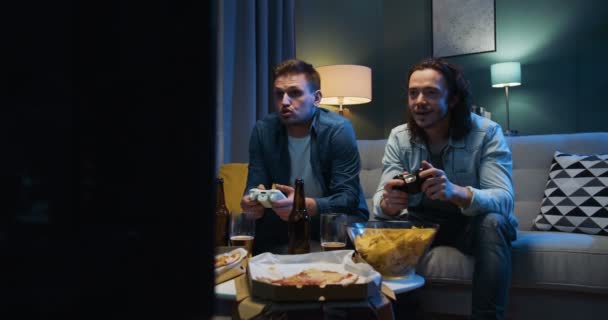 Handsome Caucasian joyful men friends sitting on sofa in tension and worrying while playing videogame with joysticks in front TV screen. Videogaming at home concept. - Imágenes, Vídeo