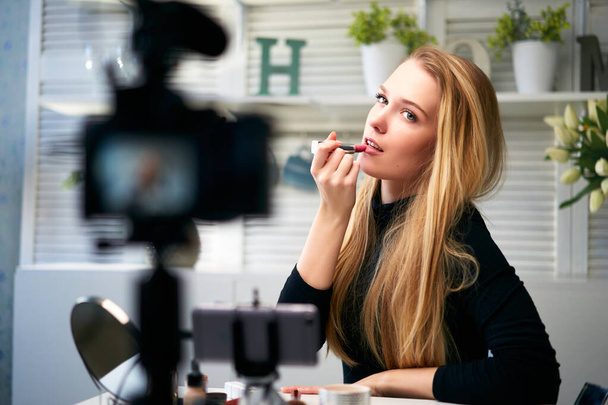 Vlogger female applies lipstick on lips. Beauty blogger woman filming daily make-up routine tutorial at camera on tripod. Influencer blonde girl live streaming cosmetics product review in home studio - Fotografie, Obrázek