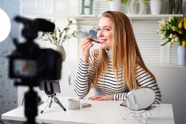 Vlogger female applies lipstick on lips. Beauty blogger woman filming daily make-up routine tutorial at camera on tripod. Influencer blonde girl live streaming cosmetics product review in home studio - Photo, Image