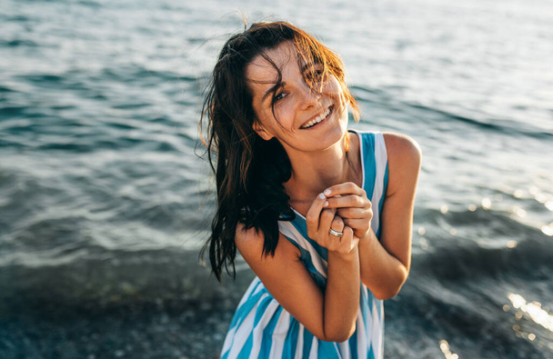 日没の背景に海の上にポーズ風吹いて髪を持つ幸せなブルネットの美しい女性の水平画像。暖かい日差しを楽しむビーチ休暇で幸せな笑顔白人の美しい女の子.  - 写真・画像