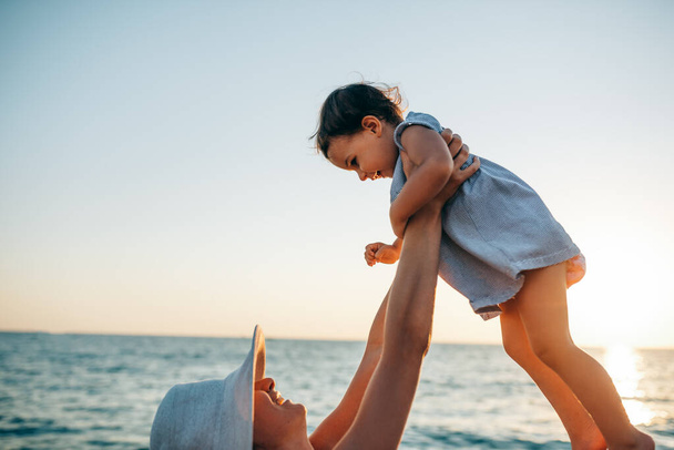 Horizontal cropped image of happy young mother playing with her daughter at the sea sunset. Smiling female with baby walking outside at the ocean beach on sunset. Motherhood love care. - 写真・画像