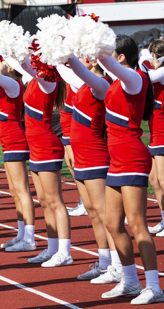 Side view of high school cheerleaders holding their pom poms up over head while cheering at their homecoming football game. - Photo, Image