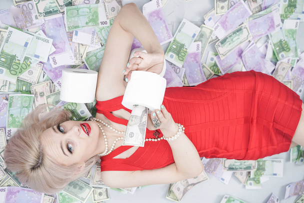 uma menina loira emocional em um vestido vermelho está deitado em dinheiro espalhado com papel higiênico. uma pessoa rica durante uma epidemia está pronta para ficar em casa e pronta para o auto-isolamento
 - Foto, Imagem
