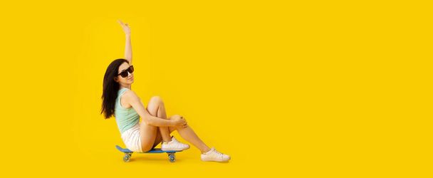 Stüdyoda güneş gözlüklü genç bir kızın sarı renkli arka planda kaykay üzerinde oturması panoramik bir görüntü. - Fotoğraf, Görsel