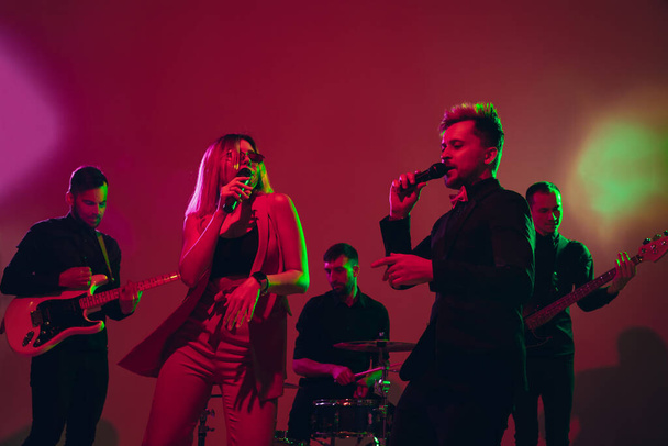 Young caucasian musicians, band performing in neon light on red studio background - Φωτογραφία, εικόνα