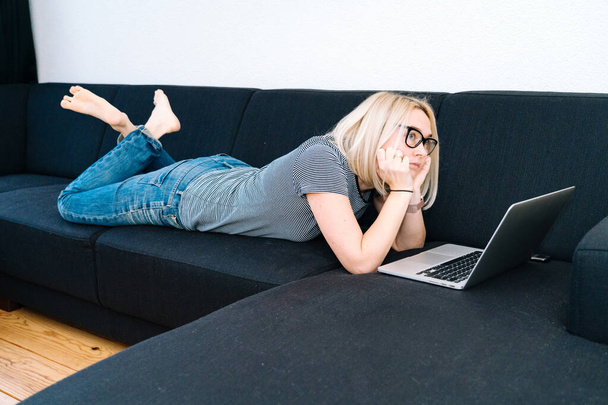 Fernunterricht und Arbeit im Online-Bereich. Büroarbeiterinnen arbeiten fernab von zu Hause. Lässige Frau mit blonden Haaren und Brille liegt mit Laptop im Wohnzimmer auf schwarzem Sofa - Foto, Bild