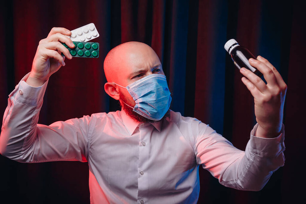 ガーゼマスクと白いシャツに身を包んだ男が黒を背景に薬や混合物を手に持っている。医療,呼吸器疾患予防,ウイルス感染症の予防, COVID-19の概念. - 写真・画像