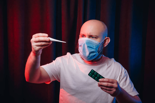 Gazlı bez maskeli, elinde termometre ve ilaç tutan, ateş belirtilerini kontrol eden bir adam. Korku ve şüphe duygusu. Sağlık, solunum hastalıklarının önlenmesi, virüs enfeksiyonlarının önlenmesi, COVID-19 konsepti. - Fotoğraf, Görsel