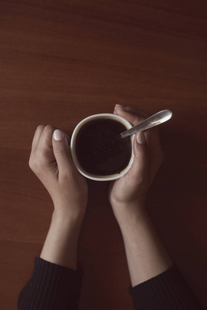 Γυναίκα στην καφετέρια. Μια αγνώριστη γυναίκα που πίνει καφέ στο άνετο περιβάλλον του σπιτιού. Γυναικεία χέρια κρατώντας φλιτζάνι καφέ σε ξύλινο τραπέζι φόντο - Φωτογραφία, εικόνα