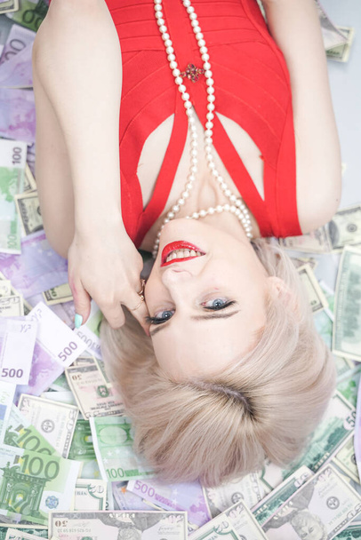 a beautiful blonde woman in a red dress is lying on a lot of money and enjoying life. rich caucasian girl top view - 写真・画像