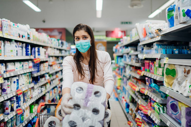 Γυναίκα αγοραστής με μάσκα και γάντια πανικού αγορά και αποθήκευση χαρτιού τουαλέτας στο κατάστημα.Προετοιμασία για πανδημία ιού παθογόνου.Prepper αγορά χύμα προμήθειες καθαρισμού λόγω Covid-19. - Φωτογραφία, εικόνα