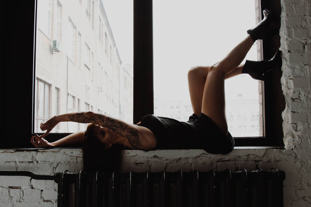 Beautiful young woman in little black dress lying on windowsill - Фото, изображение