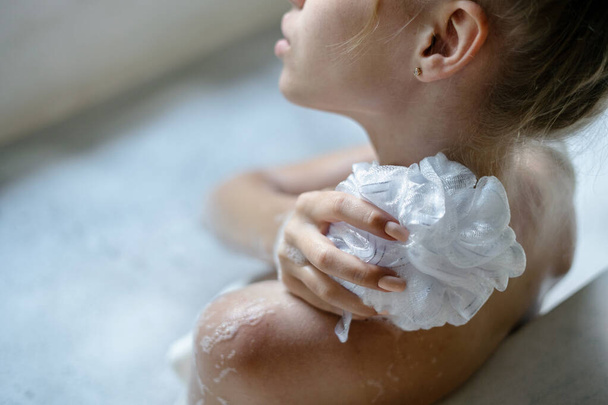 Έννοια φροντίδας σώματος. Καλλιεργημένη και υψηλής γωνίας άποψη του νεαρού ενήλικου κοριτσιού που κάνει μπάνιο, απολαμβάνοντας σαπουνόνερο αφρού, κρατώντας σφουγγάρι στο χέρι, πλένονται πίσω - Φωτογραφία, εικόνα