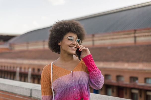 Mignonne fille multiethnique en vêtements colorés souriant parle sur son téléphone mobile dans la rue un après-midi de printemps
 - Photo, image