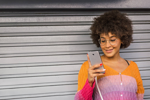 Piękna wielonarodowa dziewczyna z afro włosy ubrane w wiosennym kolorze koszuli uśmiecha się patrząc na jej smartfona. Obraz z copyspace - Zdjęcie, obraz