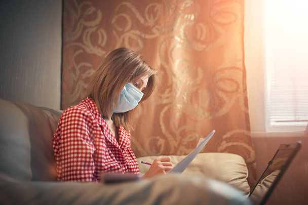 Young businesswoman in a medical protective mask works from home at the computer during self-isolation and quarantine. Virus coronavirus outbreak, flu epidemic and covid ncov novel. Stay home - Zdjęcie, obraz