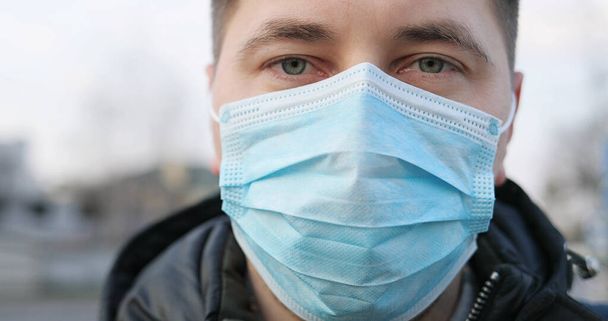 Close up of Caucasian sad ill man in medical mask standing at street and looking to camera. Portrait of guy wearing protection from coronavirus in city. Pandemic concept. Protected person at fresh air - Fotografie, Obrázek