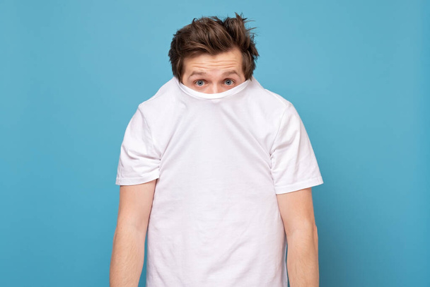 Shy caucasian man hiding behind his white shirt being afraid of coronavirus. Studio shot on blue wall. - Foto, Imagen