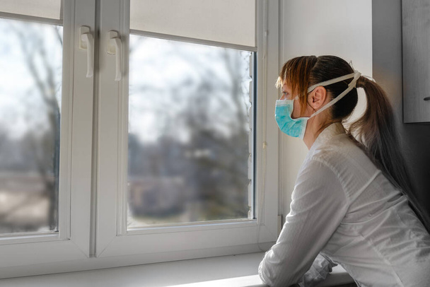 Traurige Frau mit Maske, die ins Fenster schaut. Selbstisolierung zur Verhinderung einer Pandemie des Coronavirus covid-19. - Foto, Bild