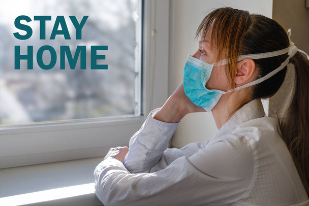 A woman wears medical mask sits in front of a window with the inscription "stay home" on a blurred city background. Stay home, call during the COVID-19 outbreak. - Photo, Image