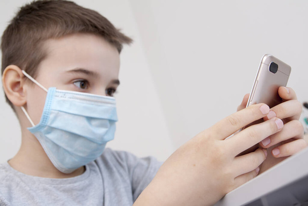 Στα χέρια ενός αγοριού ένα smartphone, μια ιατρική μάσκα στο πρόσωπό του. Καραντίνα του Coronavirus Home Schooling Concept - Φωτογραφία, εικόνα