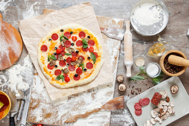 Top overhead view cooking pizza with ingredients, tomatoes, salami, flour, dough basil, spices, mozzarella and mushrooms on wooden tabletop - Foto, afbeelding