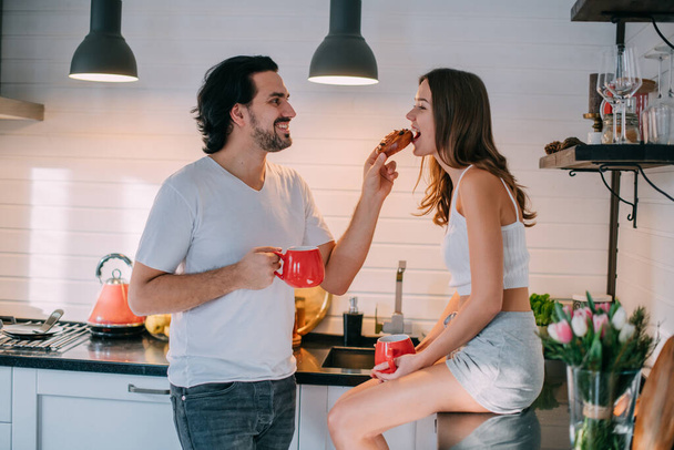 Νεαρό ζευγάρι το πρωί στην κουζίνα στο σπίτι. Άνδρας και γυναίκα έχουν ένα διασκεδαστικό πρωινό σε μια όμορφη κομψή κουζίνα. - Φωτογραφία, εικόνα