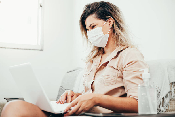 Coronavirus. Covid-19. Woman in quarantine wearing protective mask using laptop in the living room. Bottle of alcohol gel in the foreground. - Foto, afbeelding