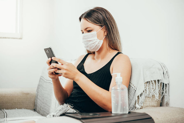 Coronavirus. Covid-19. Quarantined woman wearing protective mask making video call on cell phone. Bottle of alcohol gel in the foreground. - Photo, image