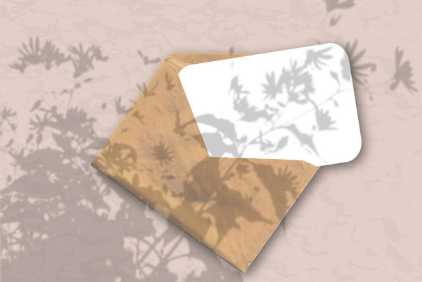 Una busta con un foglio di carta bianca testurizzata su sfondo rosa pastello. Mockup con una sovrapposizione di ombre vegetali. La luce naturale proietta ombre dalle cime di piante e fiori da campo
. - Foto, immagini