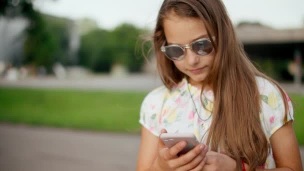 Glimlachend meisje dat buiten spelletjes speelt op de mobiele telefoon. Meisje in het park - Video