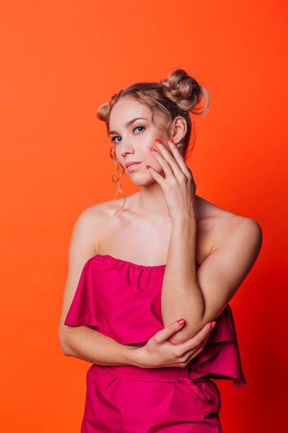 The girl in a red dress on a orange background in the studio. Blonde girl with two hair knots holding hands next to the face, looking to the camera and smiling. - Фото, зображення