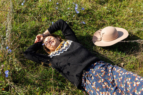 Pozowanie kobiety w naturze. Młoda kobieta w stylowych ubraniach: sweter z dzianin, kapelusz i sukienka pozowanie i odpoczynek na polu. Zdjęcia z wakacji jesienią - Zdjęcie, obraz