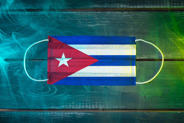 Ιατρική μάσκα προστασίας από αερομεταφερόμενες ασθένειες, βαμμένη στην εθνική σημαία της Κούβας σε μαύρο φόντο με μπλε-πράσινο καπνό. Ιατρική προστασία από αερομεταφερόμενες νόσους, coronavirus - Φωτογραφία, εικόνα