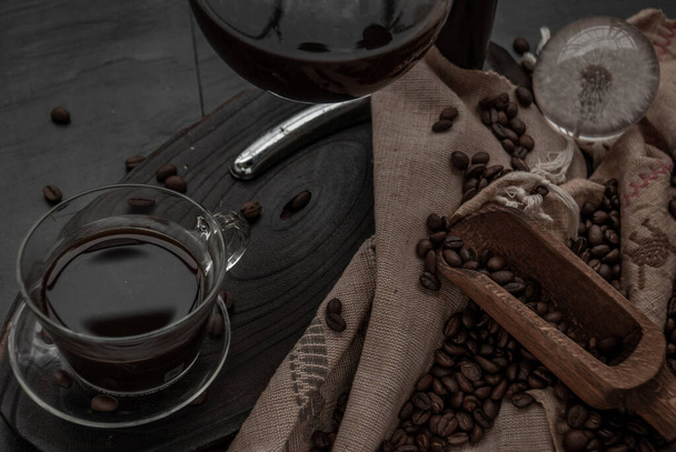 Kávový styl, šálek kávy a kávové boby, které se rozsytá starými dřevěnými naběračkami a kávovou fazolí na dřevěném a tmavém kamenném pozadí. Close-up. - Fotografie, Obrázek