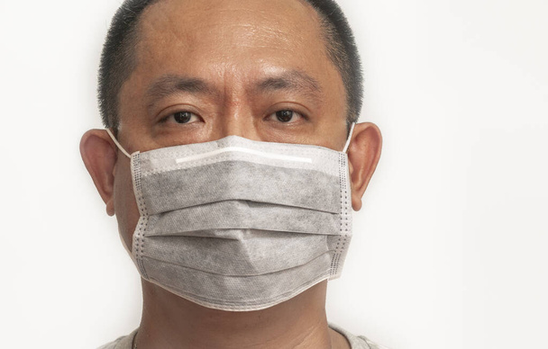 Close up face Asian middle aged man wearing medical facial mask to prevent virus or disease from the air. Isolated image on white background. Man wearing grey color mask, eyes looking at camera. - Photo, Image