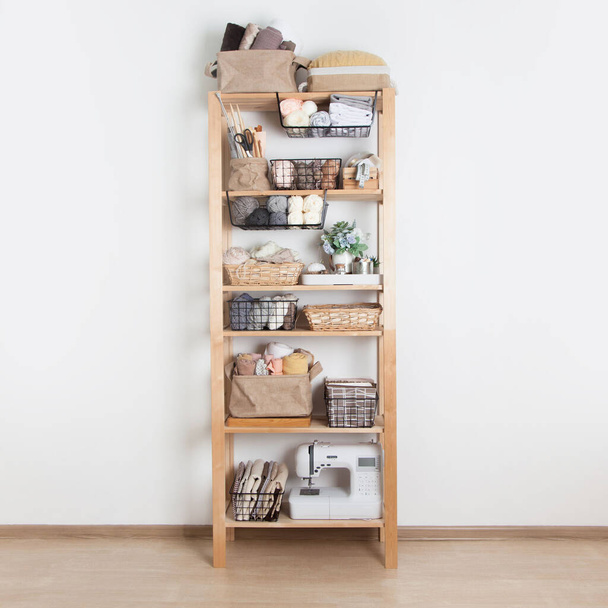 Wooden rack with baskets with a yarn cloth and sewing accessories and a sewing machine stands on a white background in a seamstress workshop. Concept of clean workplace and custom sewing. Copyspace - Zdjęcie, obraz