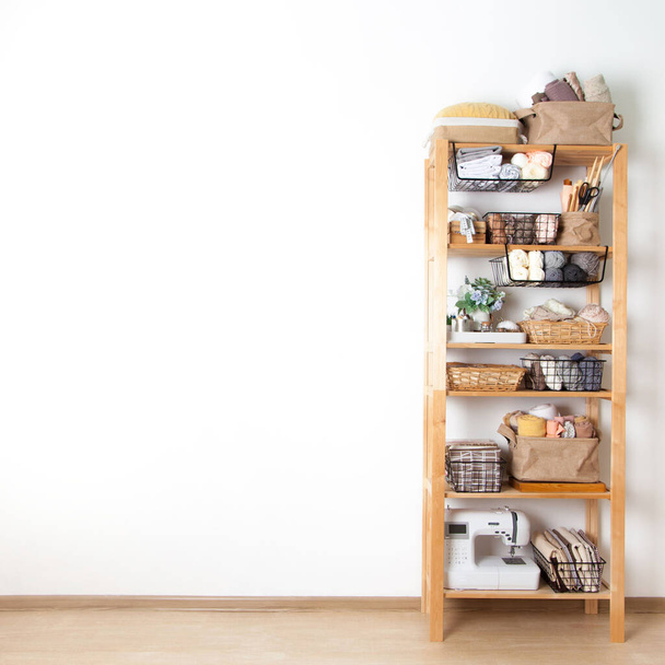 Wooden rack with baskets with a yarn cloth and sewing accessories and a sewing machine stands on a white background in a seamstress workshop. Concept of clean workplace and custom sewing. Copyspace - 写真・画像