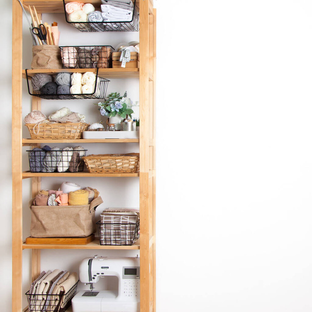 Wooden rack with baskets with a yarn cloth and sewing accessories and a sewing machine stands on a white background in a seamstress workshop. Concept of clean workplace and custom sewing. Copyspace - Фото, изображение
