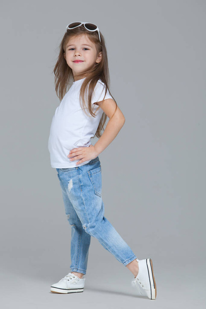 Kid girl preschooler in blue jeans, white t-shirt and sunglasses is posing isolated on grey background - Photo, image