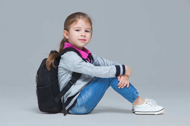 Primary school girl in jeans and uniform jacket with backpack is sitting on the floor. Isolated on grey background - Photo, image