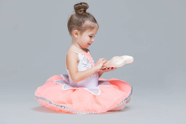 Cute adorable ballerina little girl in pink tutu sitting on the floor holding ballet shoes - Foto, Imagen
