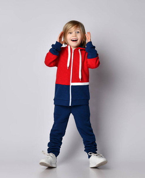 Happy excited surprised blond kid boy in red and blue sportwear with wide opened eyes holds his hand at her head - Fotografie, Obrázek