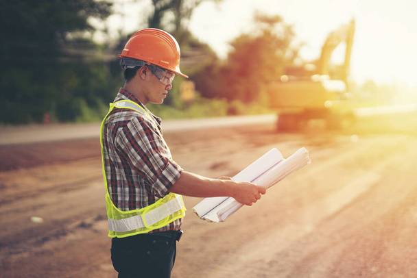 Инженер носить жесткий шлем и проведение чертеж на дорожной строительной площадке с техникой, безопасность первой концепции
 - Фото, изображение