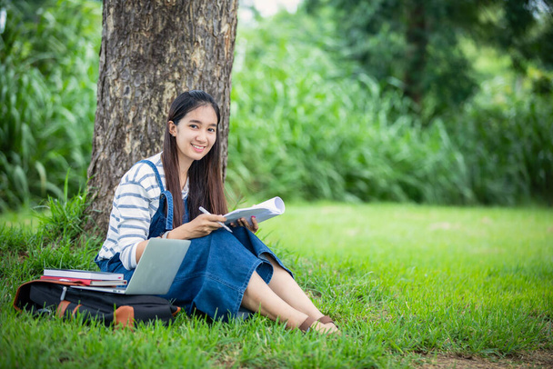 όμορφη Ασιάτισσα φοιτήτρια κρατώντας βιβλία και χαμογελώντας στην κάμερα και τη μάθηση και την εκπαίδευση έννοια στο πάρκο το καλοκαίρι για να χαλαρώσετε χρόνο - Φωτογραφία, εικόνα