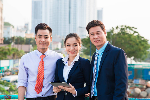Asiático hombre de negocios en traje mostrando presentación en línea en tableta digital a sus colegas mientras están de pie
 - Foto, imagen