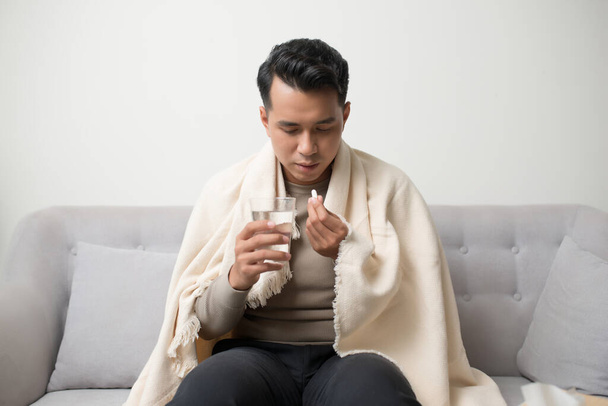 Gesundheitswesen, traditionelle Medizin und Grippekonzept - Mann nimmt Tablette gegen Kopfschmerzen auf dem Sofa sitzend. Kranker Mann behandelt Symptome einer Erkältung oder Grippe. - Foto, Bild