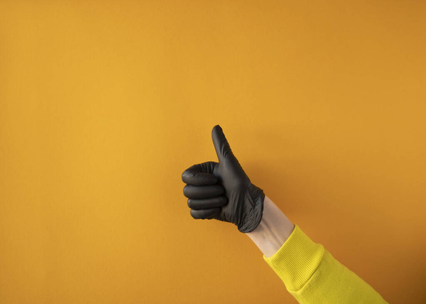 黄色の背景の黒い手袋をした女性の手の親指は - 写真・画像